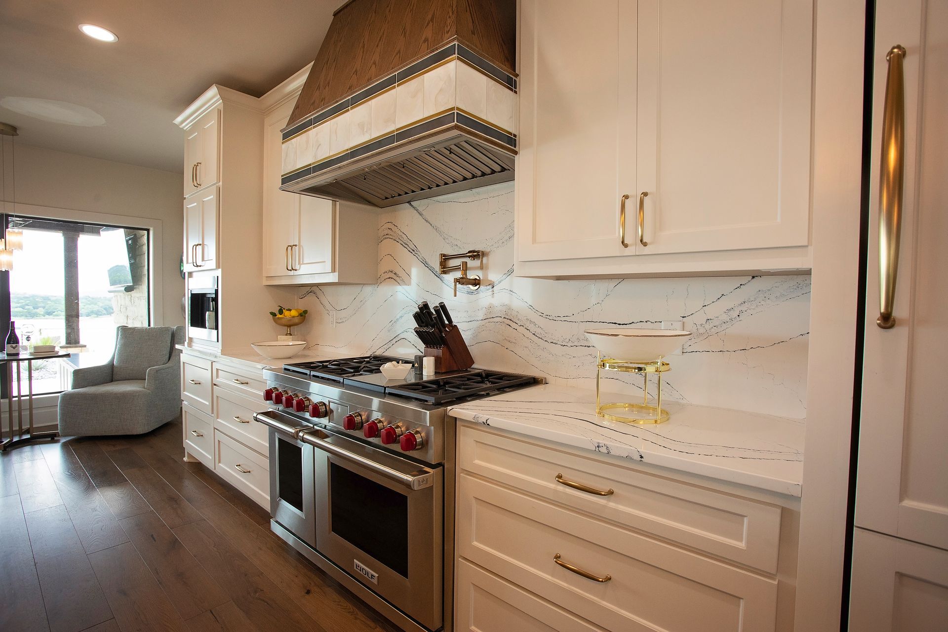 Bright modern kitchen with white cabinets, stainless stove, and a large range hood, opening into a living room.