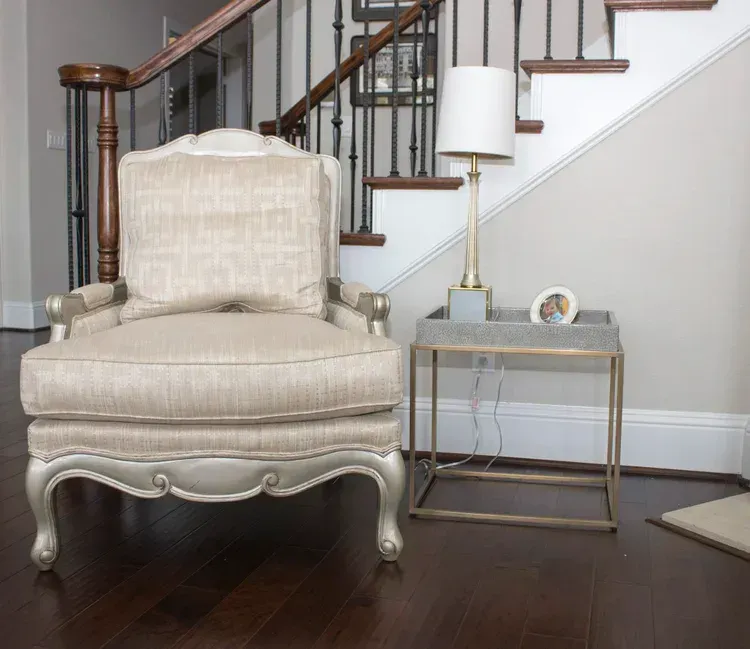 Cream upholstered armchair beside a gold side table and lamp in a stairway nook with dark wood floors
