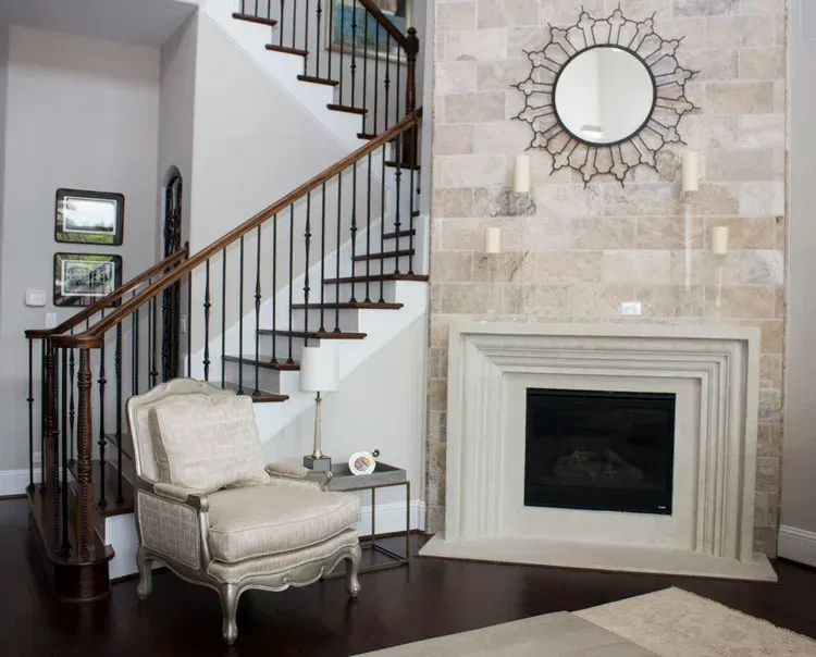 Elegant living room with staircase, cream fireplace, mirrored wall clock, and upholstered chair on dark wood floor