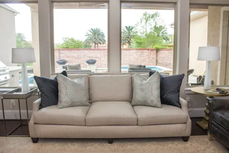 Modern living room with beige sofa, black pillows, and large windows overlooking a patio and palm trees