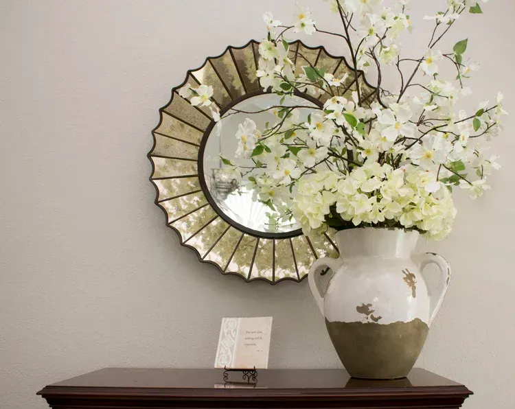 White flowers in a cream vase on a dark table beside a round mirrored wall decoration
