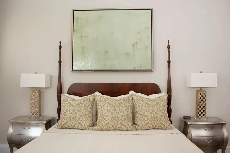 Neatly made bed with patterned pillows, white lamps, and a framed abstract painting above the headboard