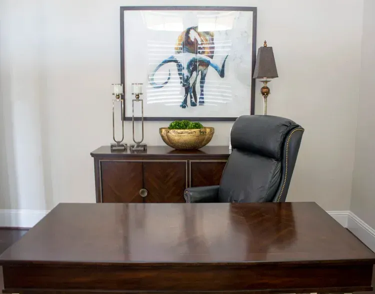 Modern office with dark wood desk, black chair, sideboard, lamp, bowl, and framed artwork on a white wall.