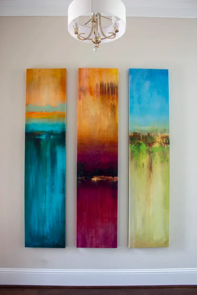 Three vertical abstract paintings in orange, turquoise, magenta, and pale green on a wall under a white chandelier