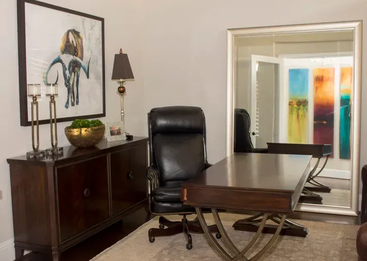 Modern office with a desk, leather chair, cabinet, lamp, and mirror in a bright room.