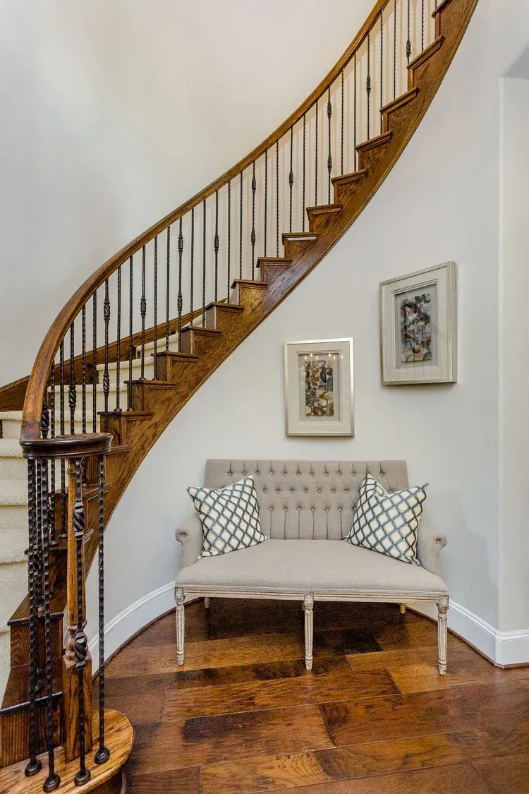 Curved wooden staircase beside a small upholstered bench on a white wall with framed art