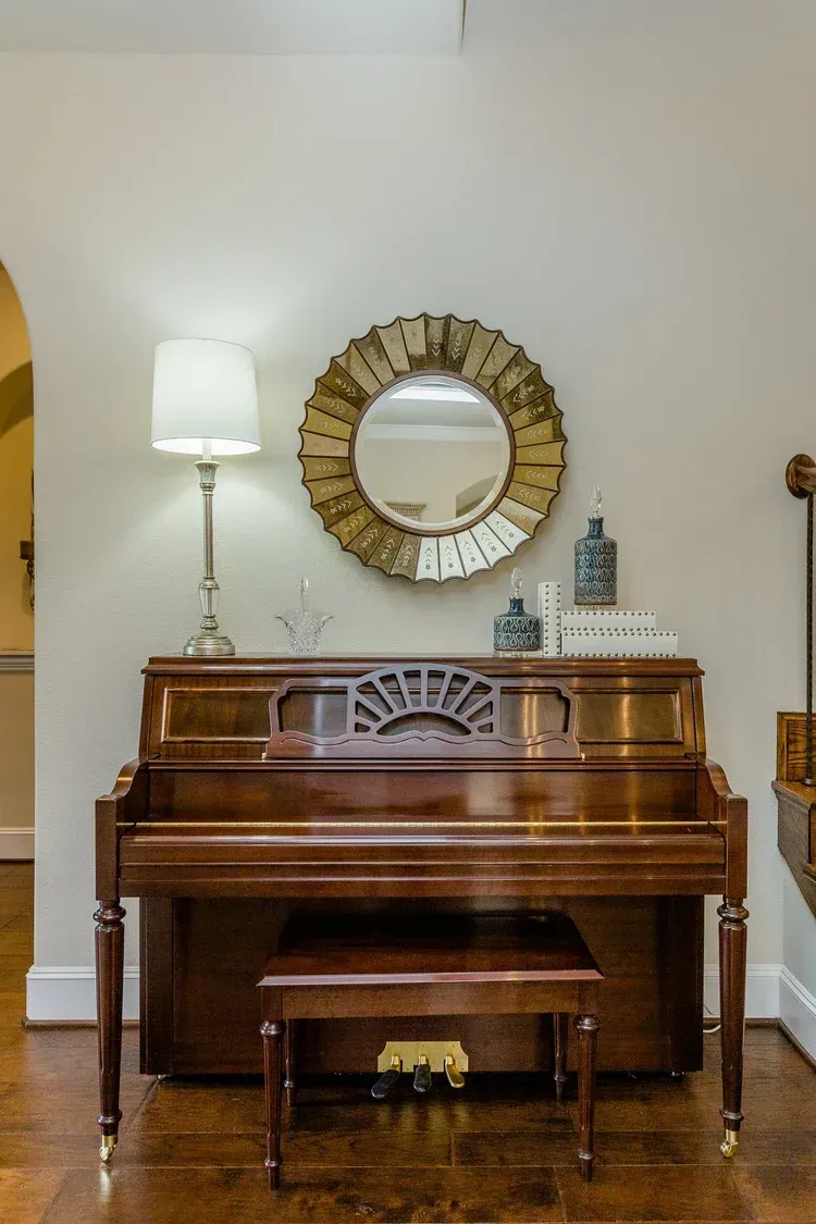 Piano against a beige wall with a round mirror, lamp, and small bench in a home interior