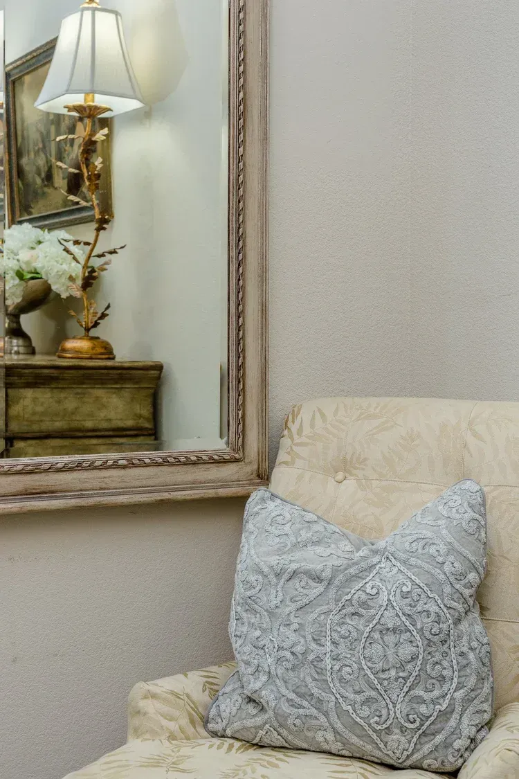Cream upholstered chair with a blue textured pillow beside a mirror and table lamp.