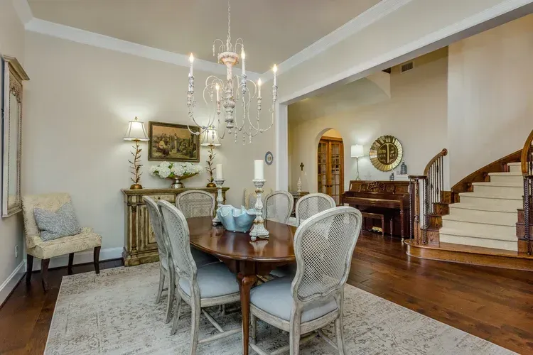 Elegant dining room with chandelier, round wooden table, upholstered chairs, staircase, and piano in the background