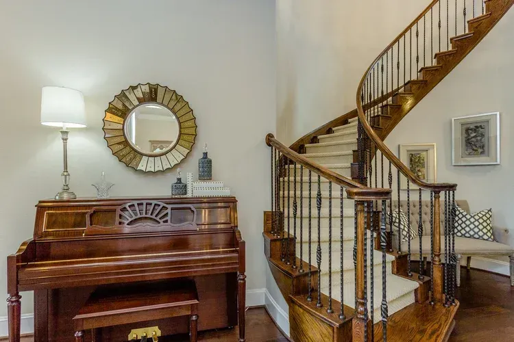 Living room with upright piano, wall mirror, lamp, and curved staircase with wooden banister