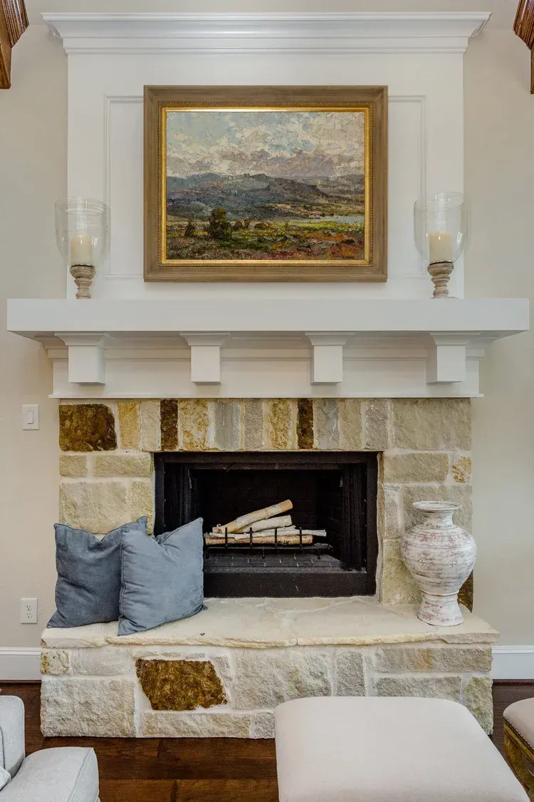 Living room fireplace with stone surround, white mantel, framed landscape art, and decorative pillows