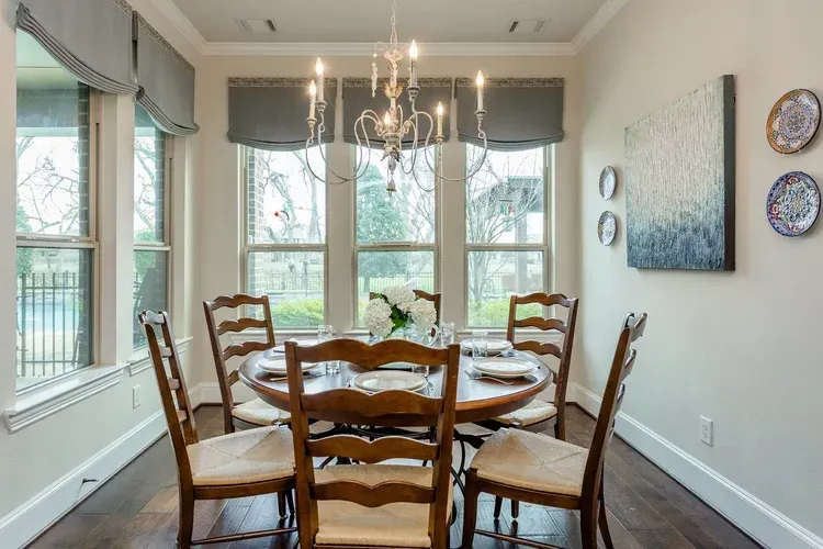 Bright dining room with a round wooden table, six chairs, chandelier, and large windows with gray shades