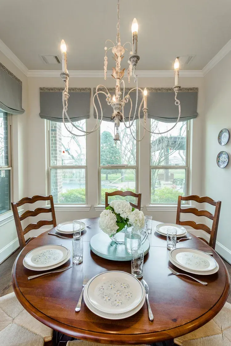 Round dining table set with white plates, glass vase centerpiece, and chandelier in a bright room.