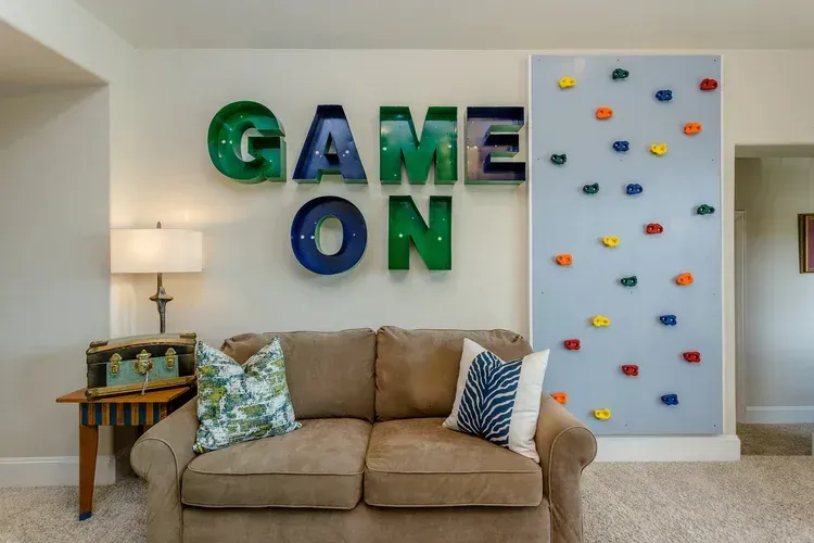 Living room with large “GAME ON” wall letters and a climbing wall panel above a beige couch