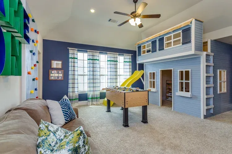 Bright playroom with a blue loft house, foosball table, sofa, and ceiling fan by a window.