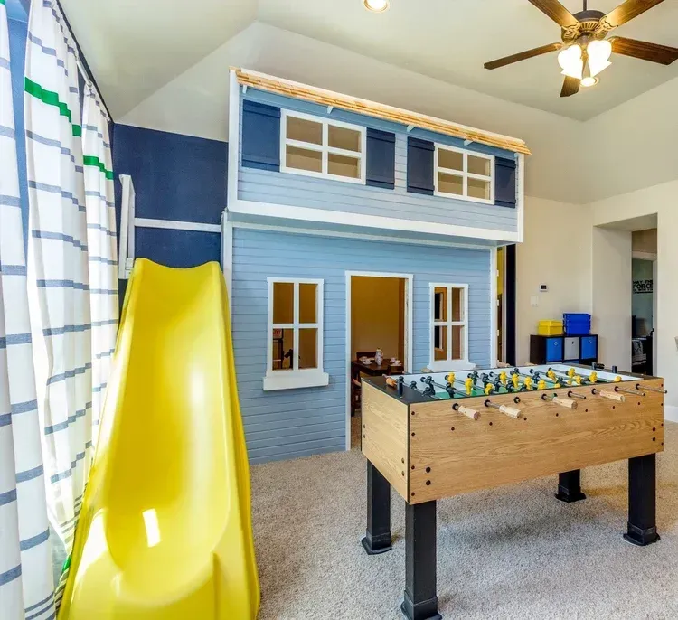 Indoor playroom with a blue playhouse, yellow slide, and foosball table beneath a ceiling fan