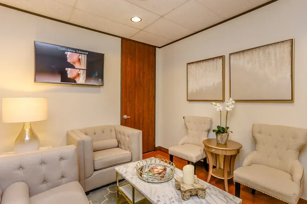 Modern waiting room with beige chairs, marble coffee table, lamp, wall TV, and orchid artwork
