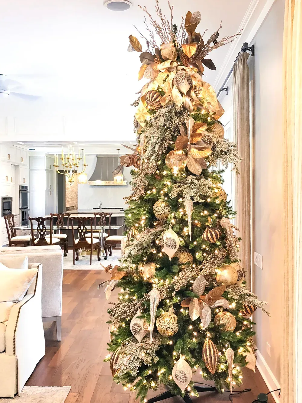 Tall decorated Christmas tree with gold ornaments and lights in a bright living room