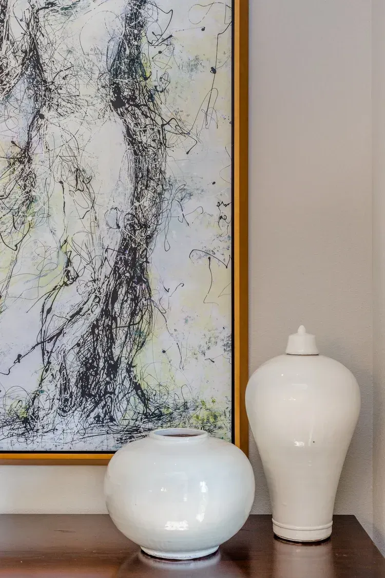 Abstract wall art beside two white ceramic vases on a shelf, with a beige wall and gold frame.
