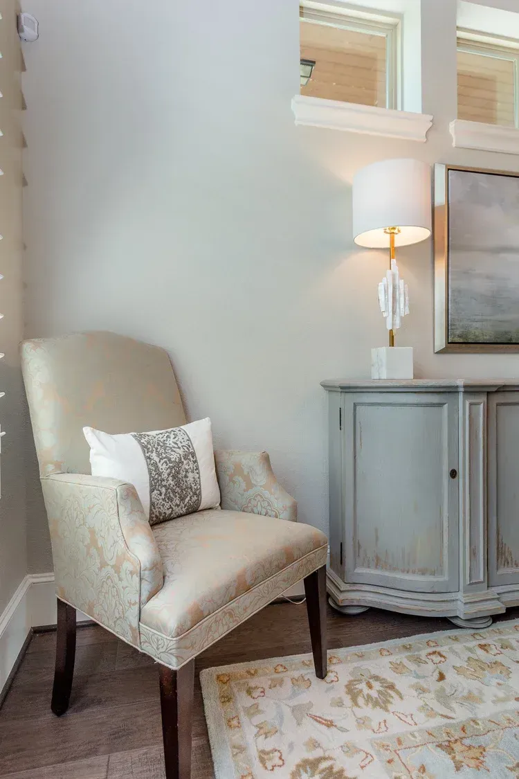 Beige armchair with patterned pillow beside a small gray side table and lamp in a bright living room corner