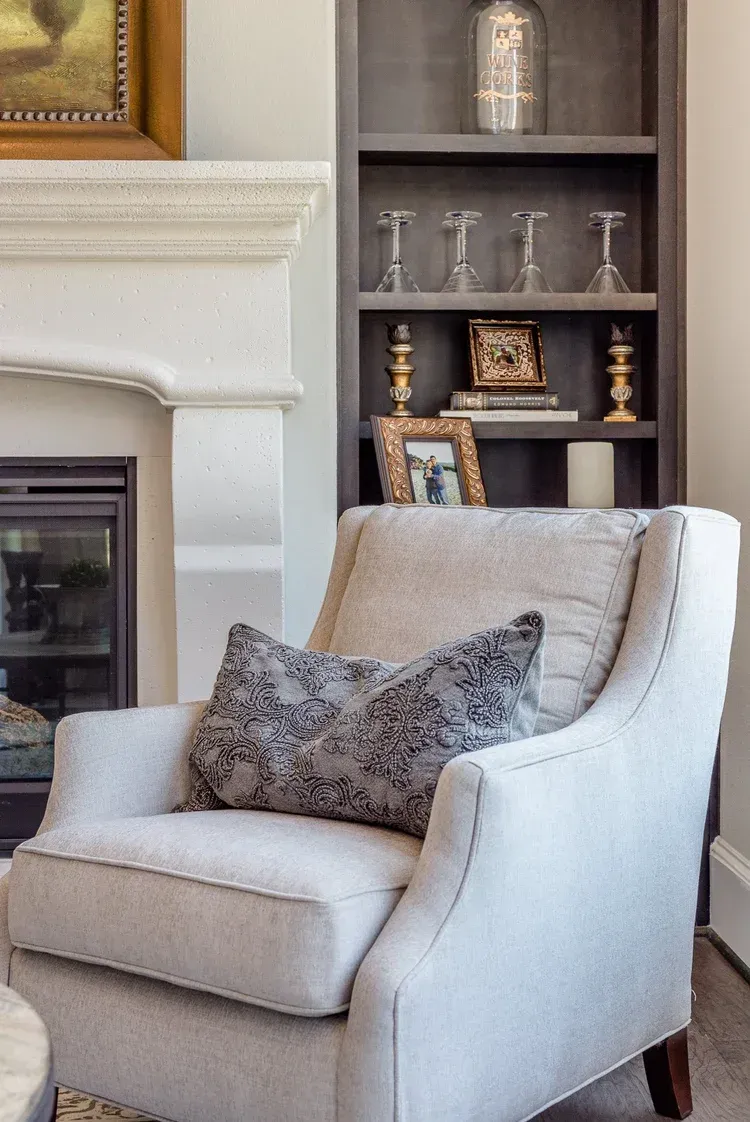 Light gray armchair with patterned pillow in a cozy living room beside a fireplace and built-in shelves