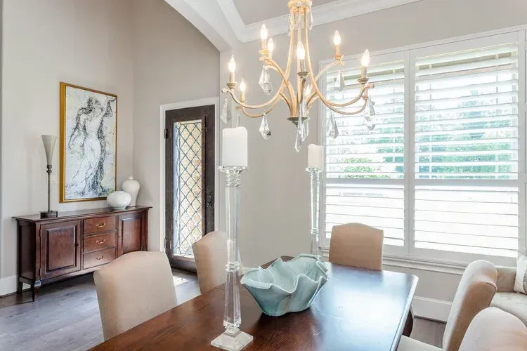 Bright dining room with chandelier, wooden table, cream chairs, sideboard, and large windows with shutters