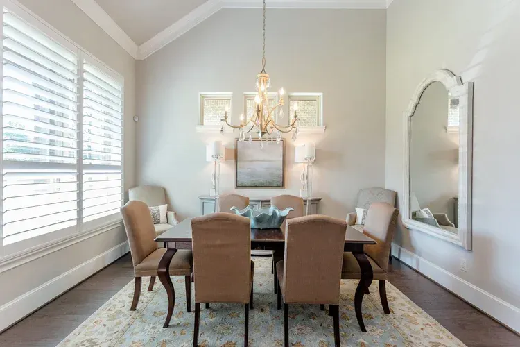 Elegant dining room with chandelier, beige chairs, large window shutters, and mirrored wall decor
