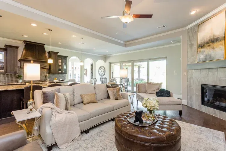 Bright open living room with beige sofa, fireplace, ottoman, and large windows facing a patio.