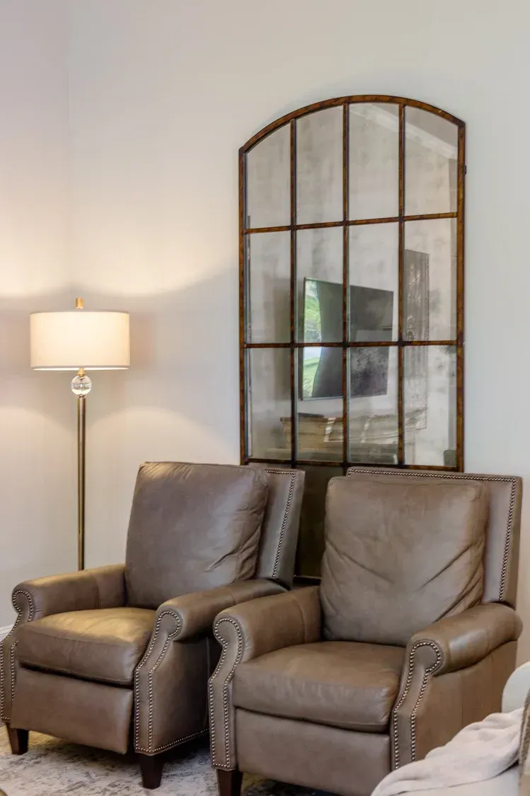 Two brown leather armchairs beside a floor lamp, with an arched mirror on a light wall.