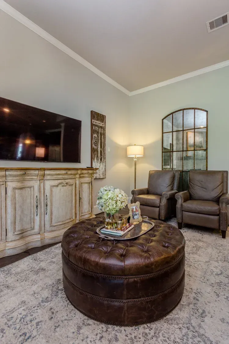 Cozy living room with TV, rustic cabinet, round ottoman, two chairs, floor lamp, and decorative mirror