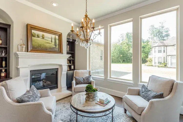 Bright living room with fireplace, chandelier, two armchairs, round coffee table, and large windows.