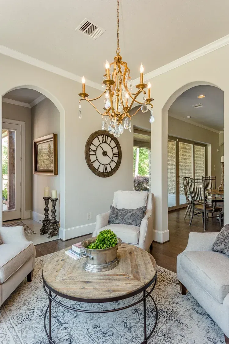 Elegant living room with chandelier, ornate clock, arched doorways, and neutral seating around a marble coffee table