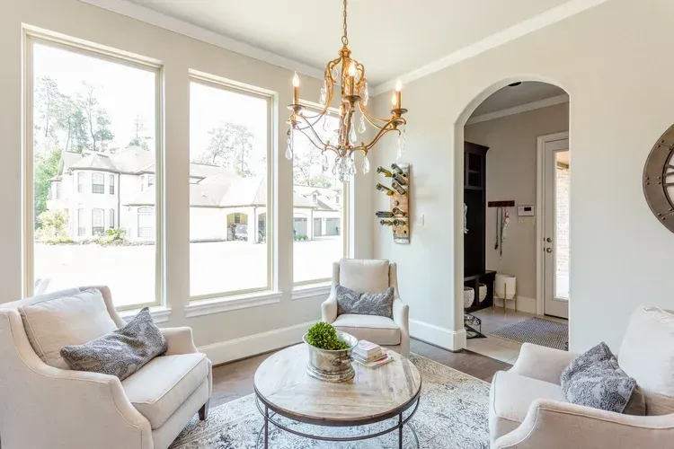 Bright living room with white chairs, glass coffee table, gold chandelier, and large windows