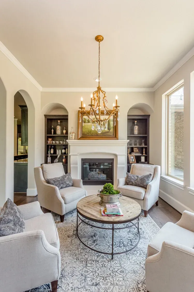 Elegant living room with white chairs, round coffee table, fireplace, chandelier, and large windows