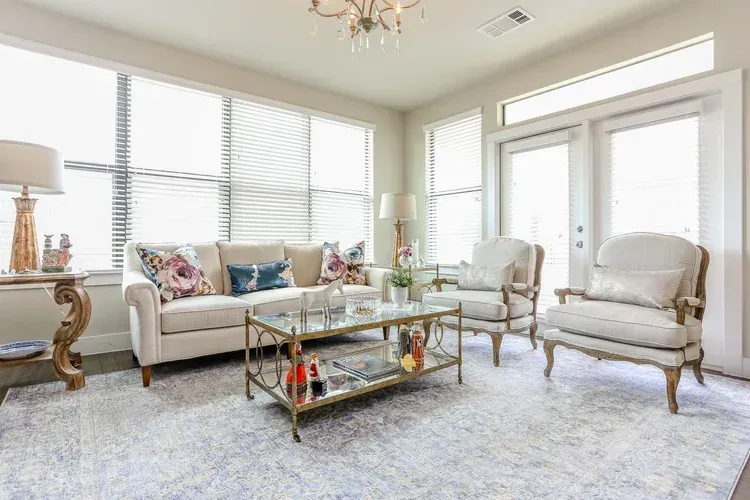 Bright living room with sofa, armchairs, glass coffee table, and large windows with white shutters