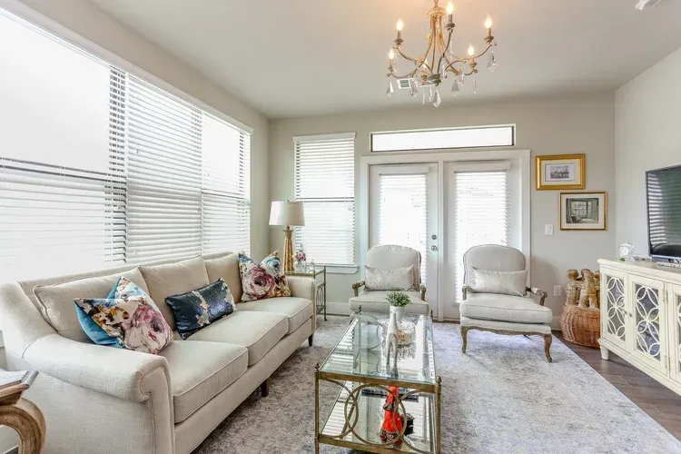 Bright living room with white sofa, two armchairs, glass coffee table, chandelier, and large windows