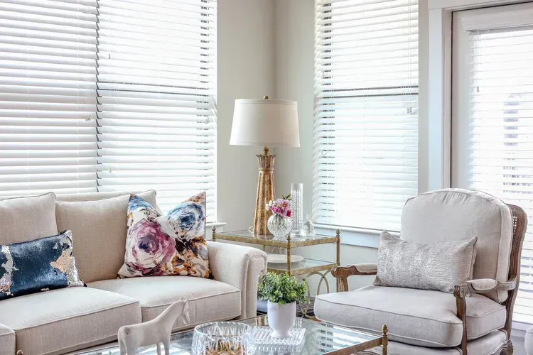 Bright living room with beige sofa, gray armchair, glass coffee table, and floral pillows by window blinds