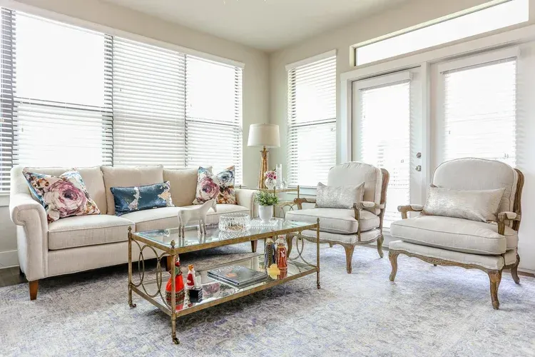 Bright living room with white sofa, two armchairs, glass coffee table, and large windows with blinds.