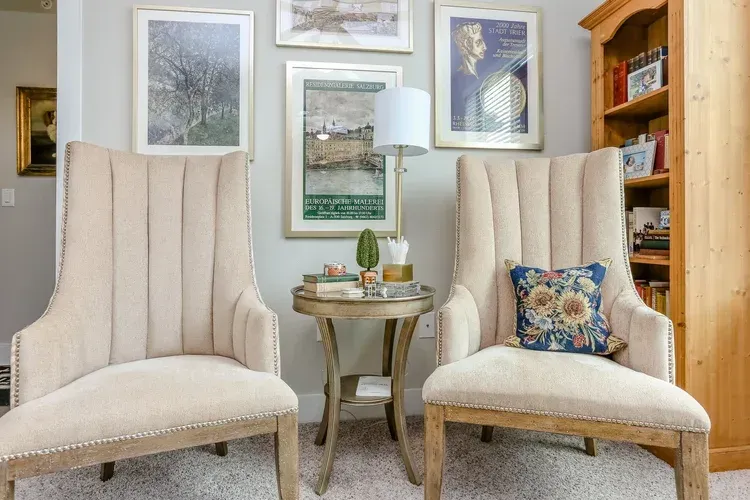Two cream wingback chairs beside a small round table in a cozy, art-filled living room