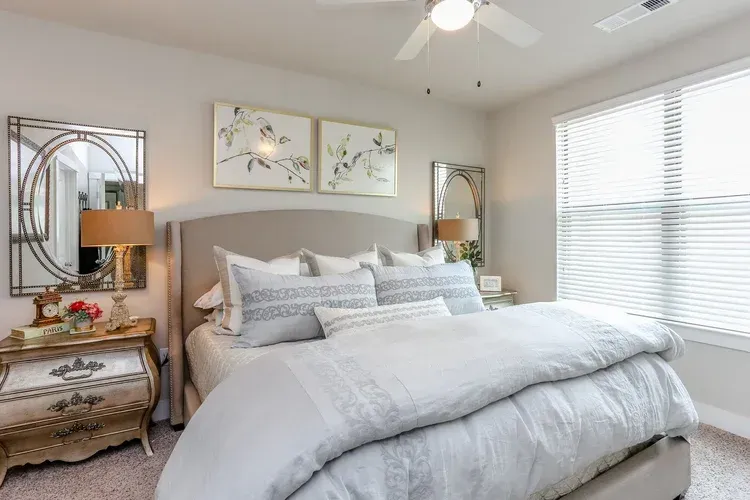 Bright bedroom with a gray bed, bedside table, wall art, mirror, and large window with blinds
