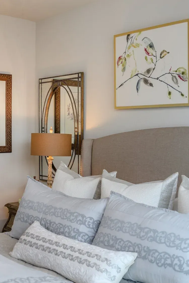 Cozy bedroom with a beige bed, patterned pillows, lamp, wall mirror, and floral artwork above the headboard