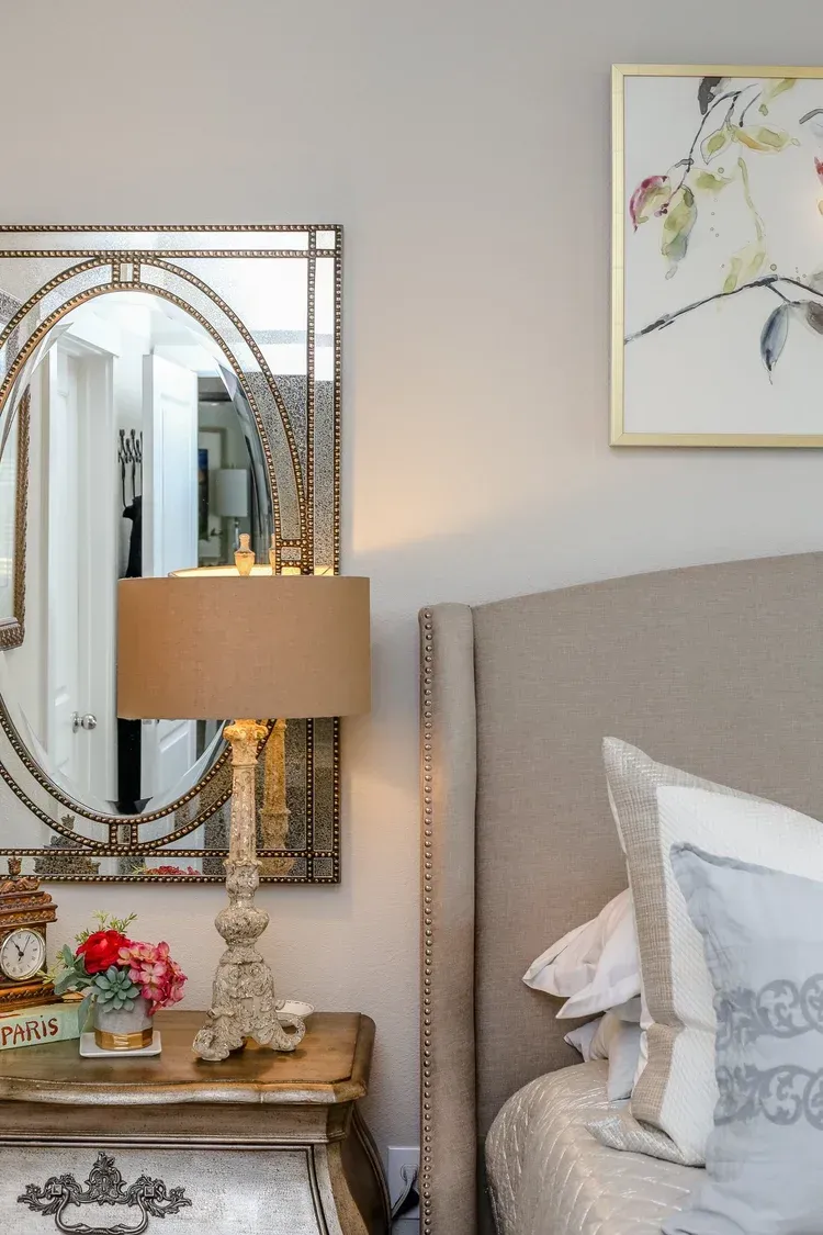 Elegant bedroom corner with upholstered bed, ornate mirror, lamp, and floral decor on a bedside table