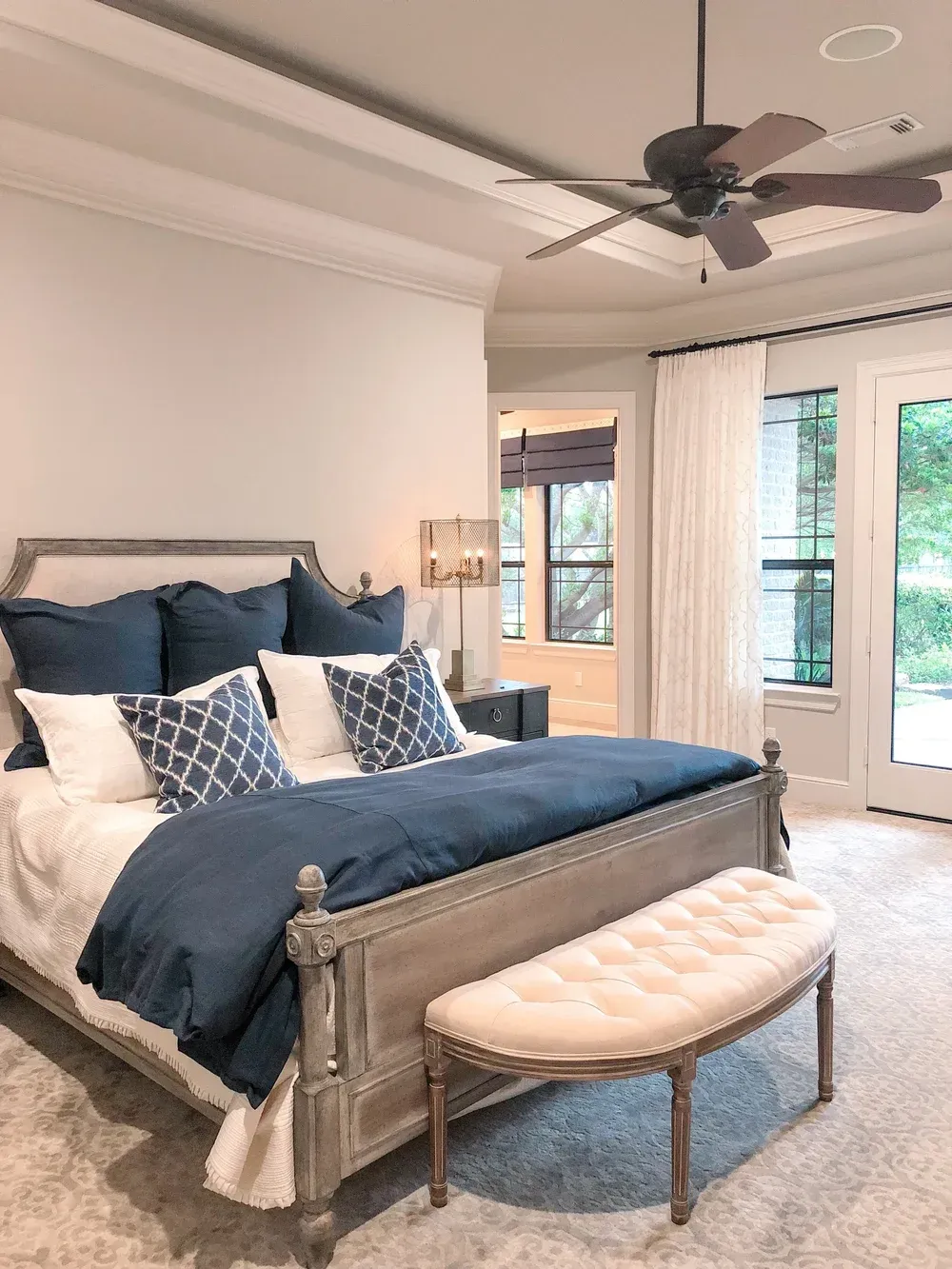 Bright bedroom with gray bed, navy bedding, tufted bench, ceiling fan, and French doors to balcony.