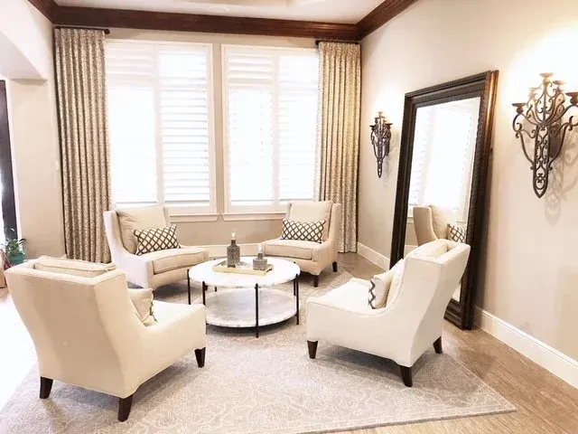 Bright sitting room with cream chairs, round table, tall mirror, and large window with curtains.