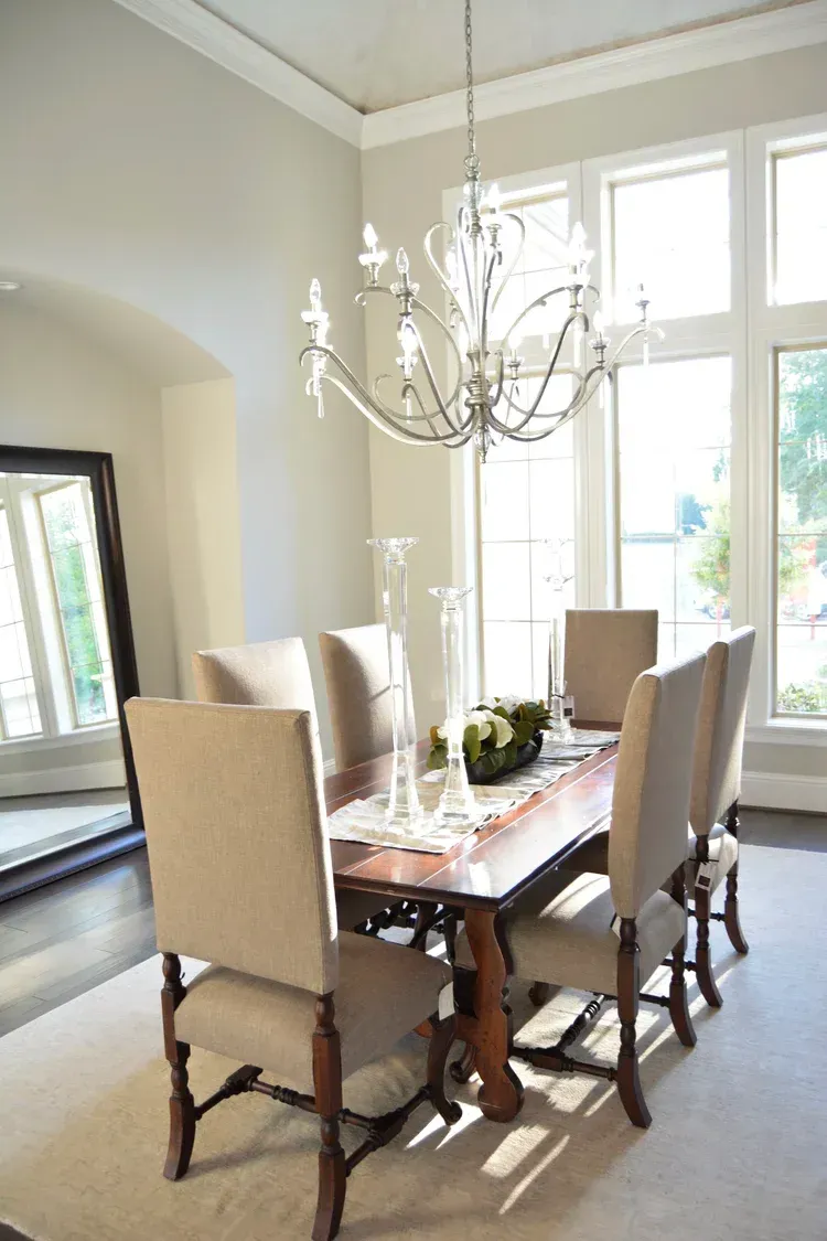Dining room with a glass table, upholstered chairs, and a chandelier by tall windows.
