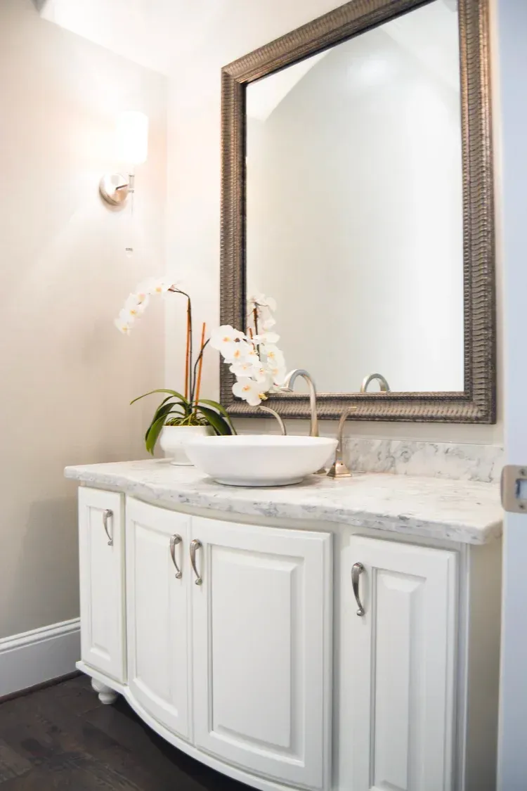 White bathroom vanity with marble countertop, vessel sink, orchid, and large framed mirror.