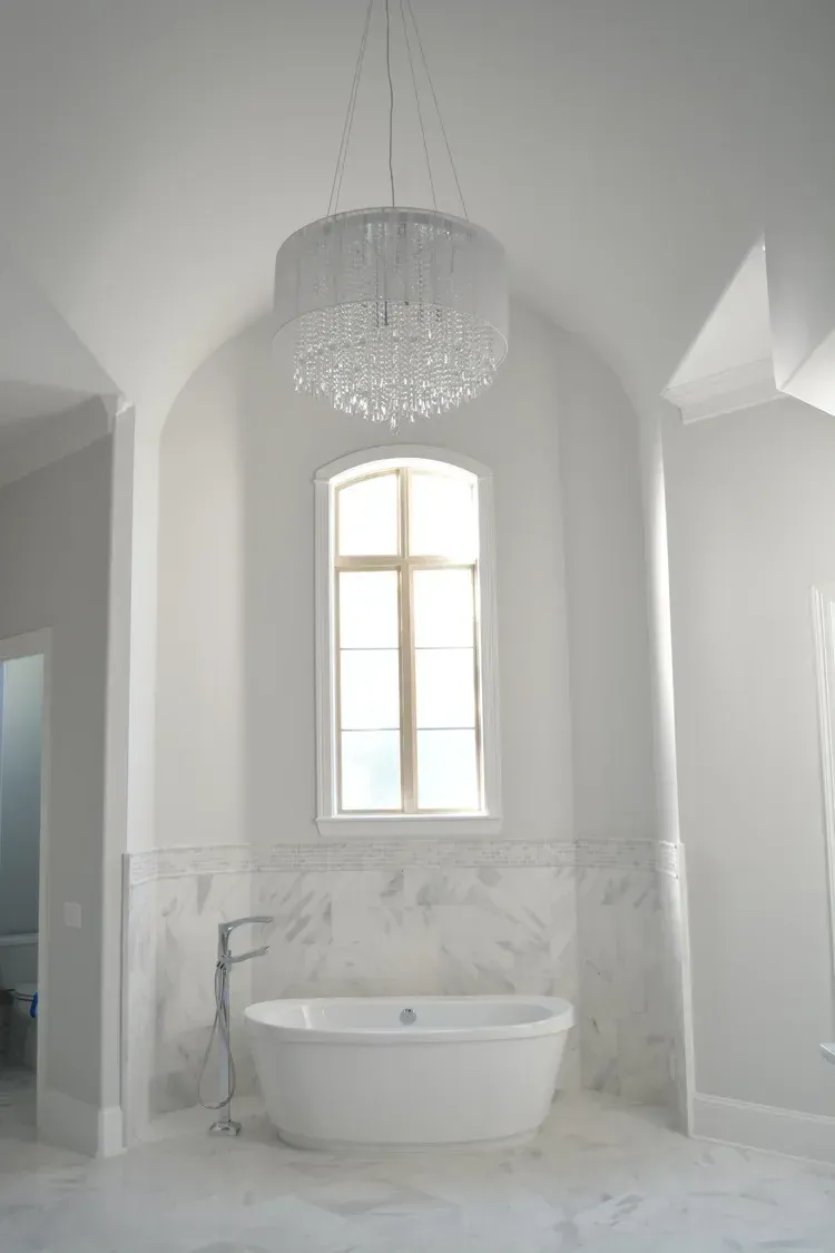 Bright white bathroom with a freestanding tub beneath a window and chandelier