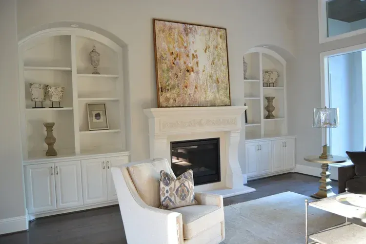 Bright living room with white built-in shelves, a fireplace, abstract art, and a cream chair by a window.