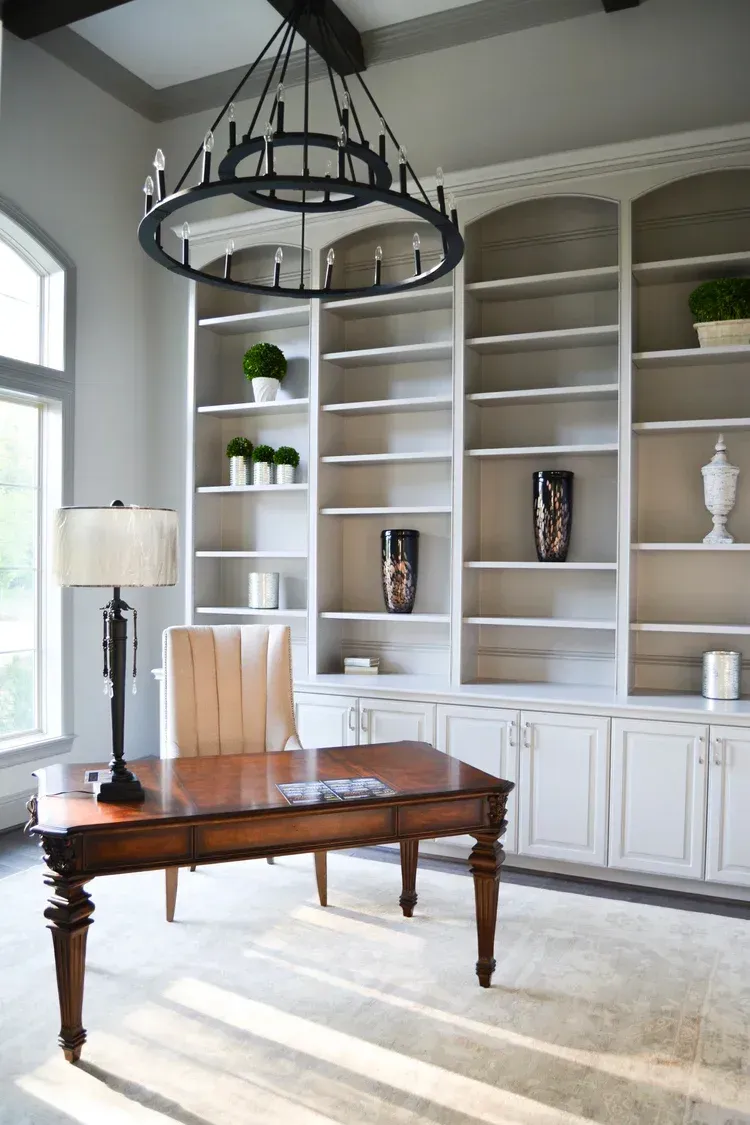 Elegant home office with built-in shelves, wooden desk, chandelier, and a large window.