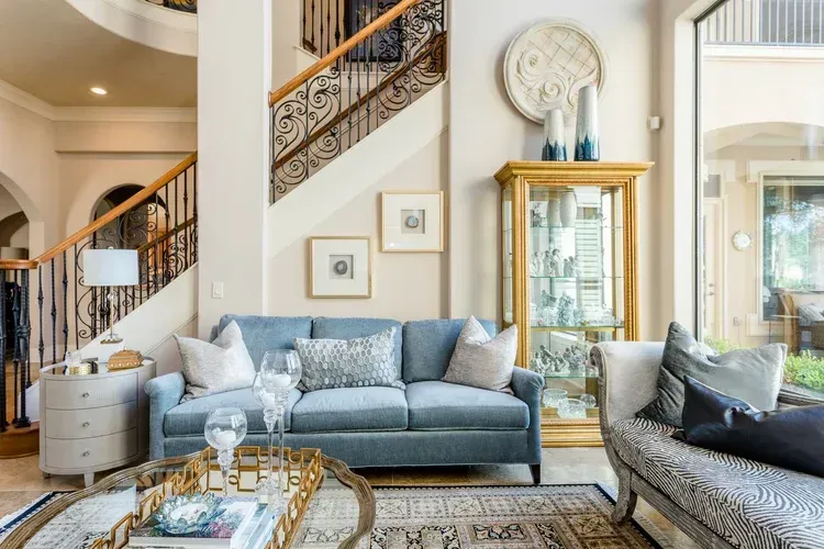 Bright living room with blue sofas, glass coffee table, staircase, and gold-framed mirror
